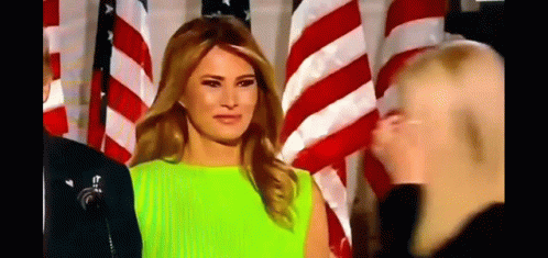 A Woman In A Neon Green Dress Is Standing In Front Of American Flags And Talking To A Man . GIF