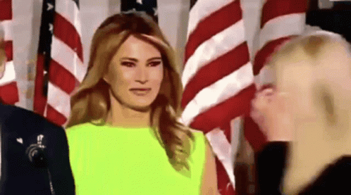 A Woman In A Neon Yellow Dress Is Standing In Front Of An American Flag . GIF