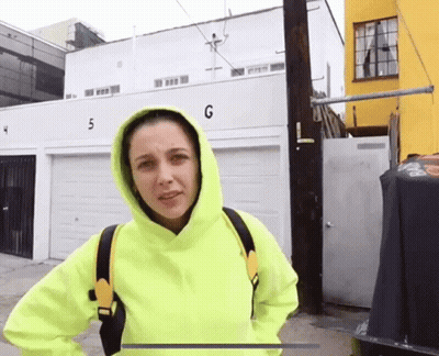 A Woman In A Neon Yellow Hoodie Stands In Front Of A Building With The Number G On It GIF
