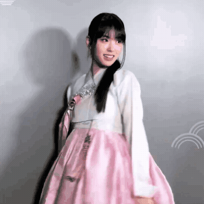 A Woman In A Pink And White Dress Is Standing In Front Of A Wall And Smiling . GIF