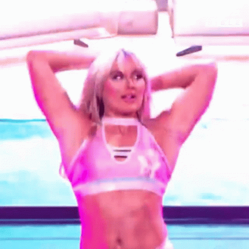 A Woman In A Pink Crop Top Is Standing On A Stage With Her Arms Behind Her Head . GIF