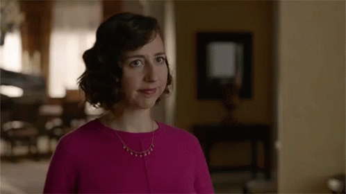 A Woman In A Pink Dress And Necklace Is Standing In A Living Room Looking At The Camera . GIF