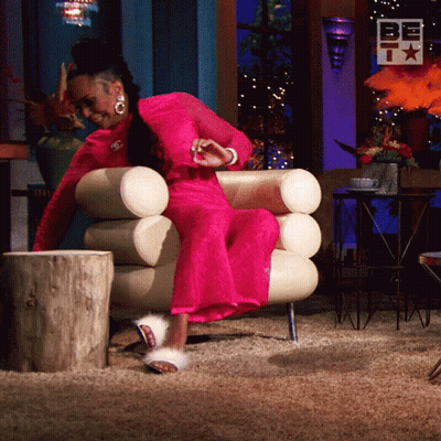 A Woman In A Pink Dress Is Sitting In A Chair With A Sign That Says Be GIF