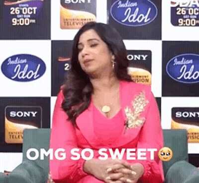 A Woman In A Pink Dress Is Sitting In Front Of A Wall With Indian Idol Logos On It GIF