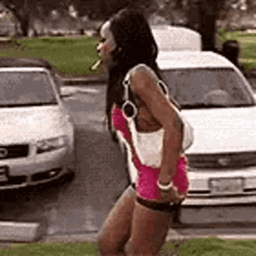 A Woman In A Pink Dress Is Smoking A Cigarette While Standing In Front Of A Parking Lot . GIF