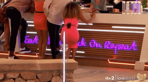A Woman In A Pink Dress Is Standing In Front Of A Neon Sign That Says " Eat Drink On Repeat " GIF