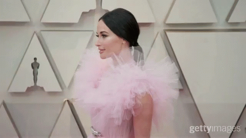A Woman In A Pink Dress Is Standing In Front Of A Wall Of Oscar Statues GIF