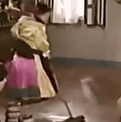 A Woman In A Pink Dress Is Standing On A Wooden Floor In A Room With A Broom . GIF