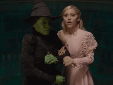 A Woman In A Pink Dress Stands Next To A Green Witch GIF