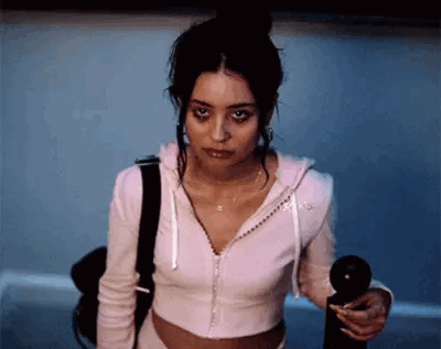 A Woman In A Pink Hoodie Is Standing In A Dark Room Holding A Black Object In Her Hand . GIF