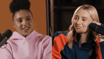 A Woman In A Pink Hoodie Is Talking Into A Microphone Next To A Woman In An Orange Jacket . GIF