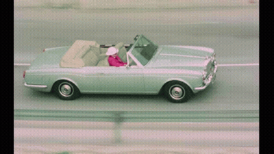 A Woman In A Pink Jacket Is Driving A Silver Convertible GIF