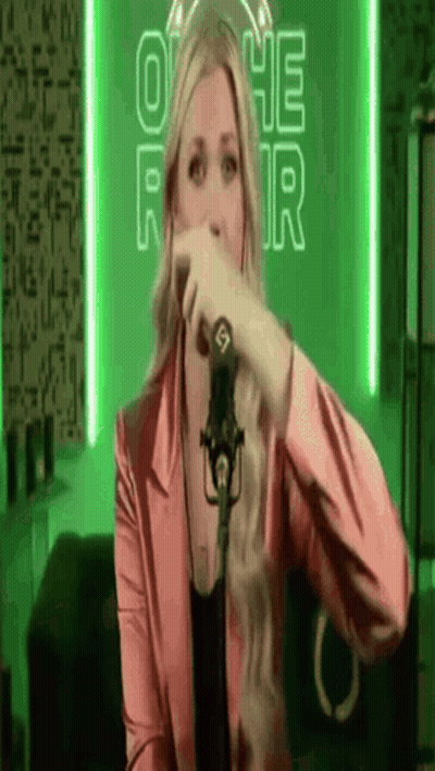 A Woman In A Pink Jacket Is Singing Into A Microphone In Front Of A Green Background . GIF