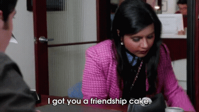 A Woman In A Pink Jacket Is Sitting At A Desk Talking To A Man And Saying I Got You A Friendship Cake GIF