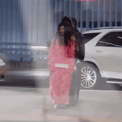 A Woman In A Pink Jacket Is Walking In Front Of A Car GIF