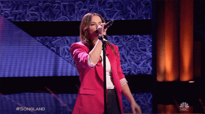 A Woman In A Pink Jacket Sings Into A Microphone On A Stage With A Nbc Logo Behind Her GIF