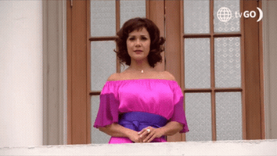 A Woman In A Pink Off The Shoulder Dress Stands On A Balcony With A Tvgo Logo In The Background GIF