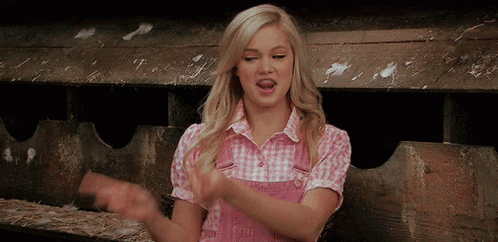 A Woman In A Pink Plaid Shirt Is Clapping Her Hands In A Barn GIF