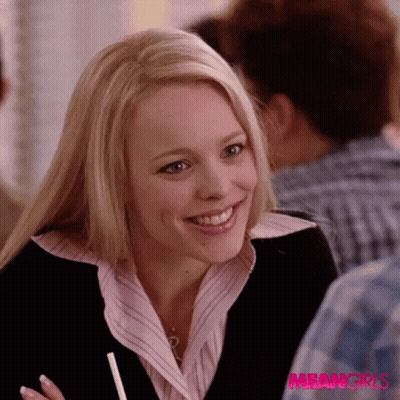 A Woman In A Pink Shirt And Black Jacket Is Smiling In Front Of A Sign That Says Meangirls GIF