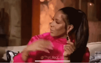 A Woman In A Pink Shirt And Earrings Is Sitting On A Couch With Her Hands On Her Chest . GIF