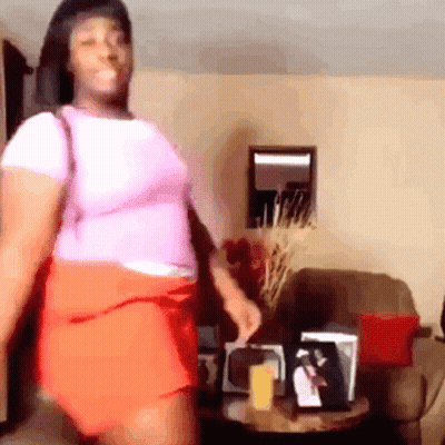 A Woman In A Pink Shirt And Orange Skirt Is Standing In A Living Room . GIF