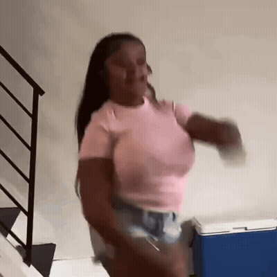 A Woman In A Pink Shirt And Shorts Is Dancing In Front Of A Blue Cooler In A Room . GIF
