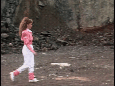 A Woman In A Pink Shirt And White Pants Walks On A Dirt Road GIF