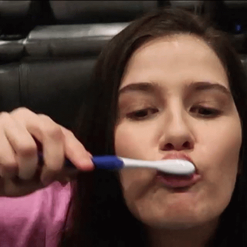 A Woman In A Pink Shirt Is Brushing Her Teeth With A Toothbrush GIF