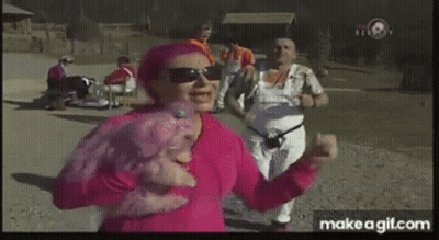 A Woman In A Pink Shirt Is Dancing In A Park With Other People . GIF