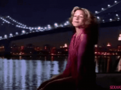 A Woman In A Pink Shirt Is Standing In Front Of A Body Of Water And A Bridge . GIF