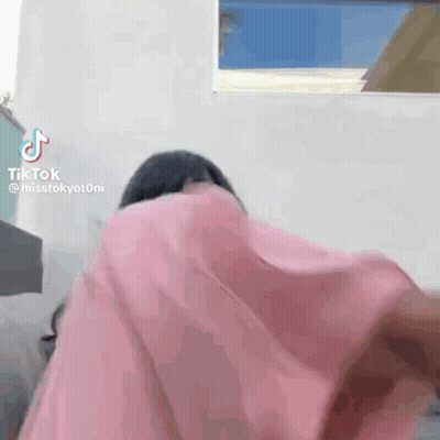 A Woman In A Pink Shirt Is Standing In Front Of A Window And Covering Her Face With A Pink Cloth . GIF