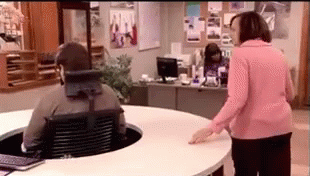 A Woman In A Pink Shirt Is Standing Next To A Man In A Wheelchair At A Desk . GIF