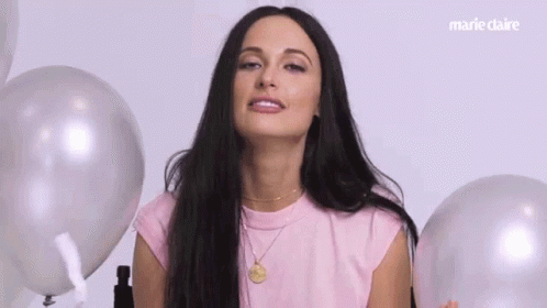 A Woman In A Pink Shirt Is Surrounded By Balloons And Making A Funny Face . GIF