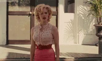 A Woman In A Pink Skirt And A White Shirt Is Standing In Front Of A Door . GIF