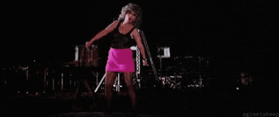 A Woman In A Pink Skirt Is Singing Into A Microphone On Stage . GIF