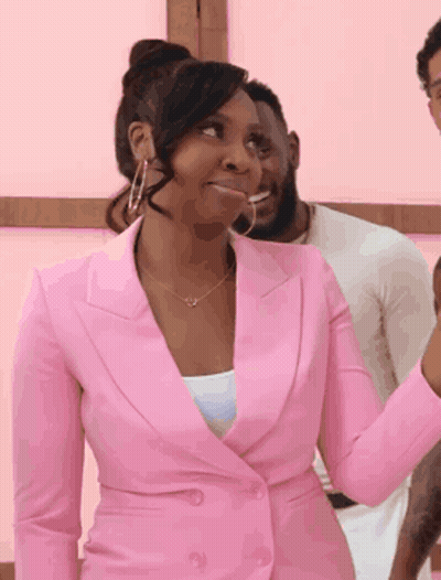 A Woman In A Pink Suit And Hoop Earrings Is Standing In Front Of A Man . GIF