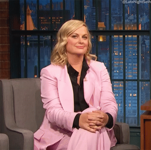 A Woman In A Pink Suit Is Sitting In A Chair With Her Legs Crossed And Her Hands Folded . GIF