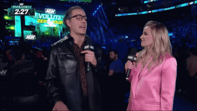 A Woman In A Pink Suit Is Talking To A Man In A Leather Jacket With A Microphone In Front Of An Aew Sign GIF