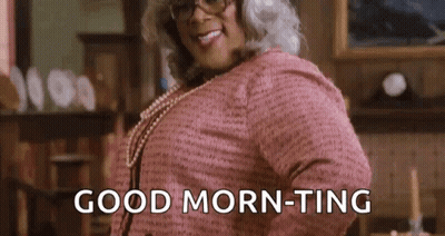 A Woman In A Pink Sweater And Pearls Is Standing In A Living Room And Saying `` Good Morning '' . GIF