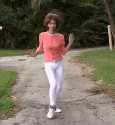 A Woman In A Pink Sweater And White Pants Is Walking Down A Sidewalk . GIF