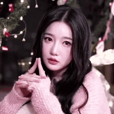 A Woman In A Pink Sweater Is Sitting In Front Of A Christmas Tree With Her Hands Folded . GIF