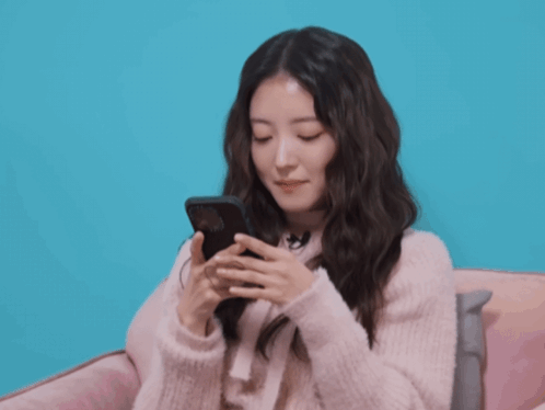 A Woman In A Pink Sweater Is Smiling While Holding A Cell Phone GIF