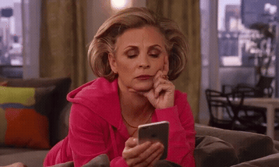 A Woman In A Pink Sweatshirt Is Laying On A Couch Looking At Her Phone . GIF