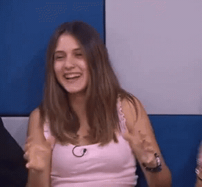 A Woman In A Pink Tank Top Is Sitting In Front Of A Blue Wall And Laughing . GIF
