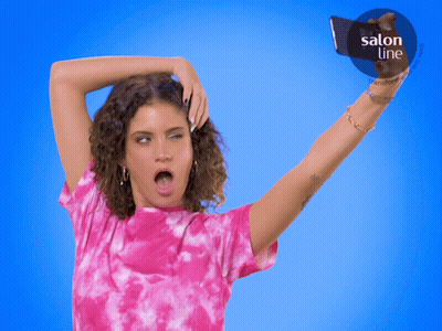 A Woman In A Pink Tie Dye Shirt Takes A Selfie In Front Of A Blue Background That Says Salon Line GIF