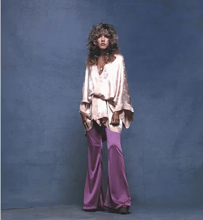 A Woman In A Pink Top And Purple Pants Stands In Front Of A Blue Background GIF