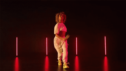 A Woman In A Pink Top And White Pants Is Dancing On A Dark Stage GIF