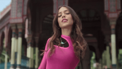A Woman In A Pink Top Is Standing In Front Of A Building With Her Eyes Closed GIF