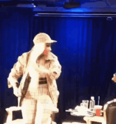 A Woman In A Plaid Coat And Hat Is Standing On A Stage . GIF