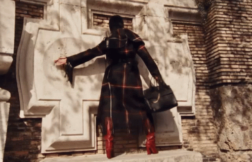 A Woman In A Plaid Coat And Red Boots Is Standing In Front Of A Brick Building GIF
